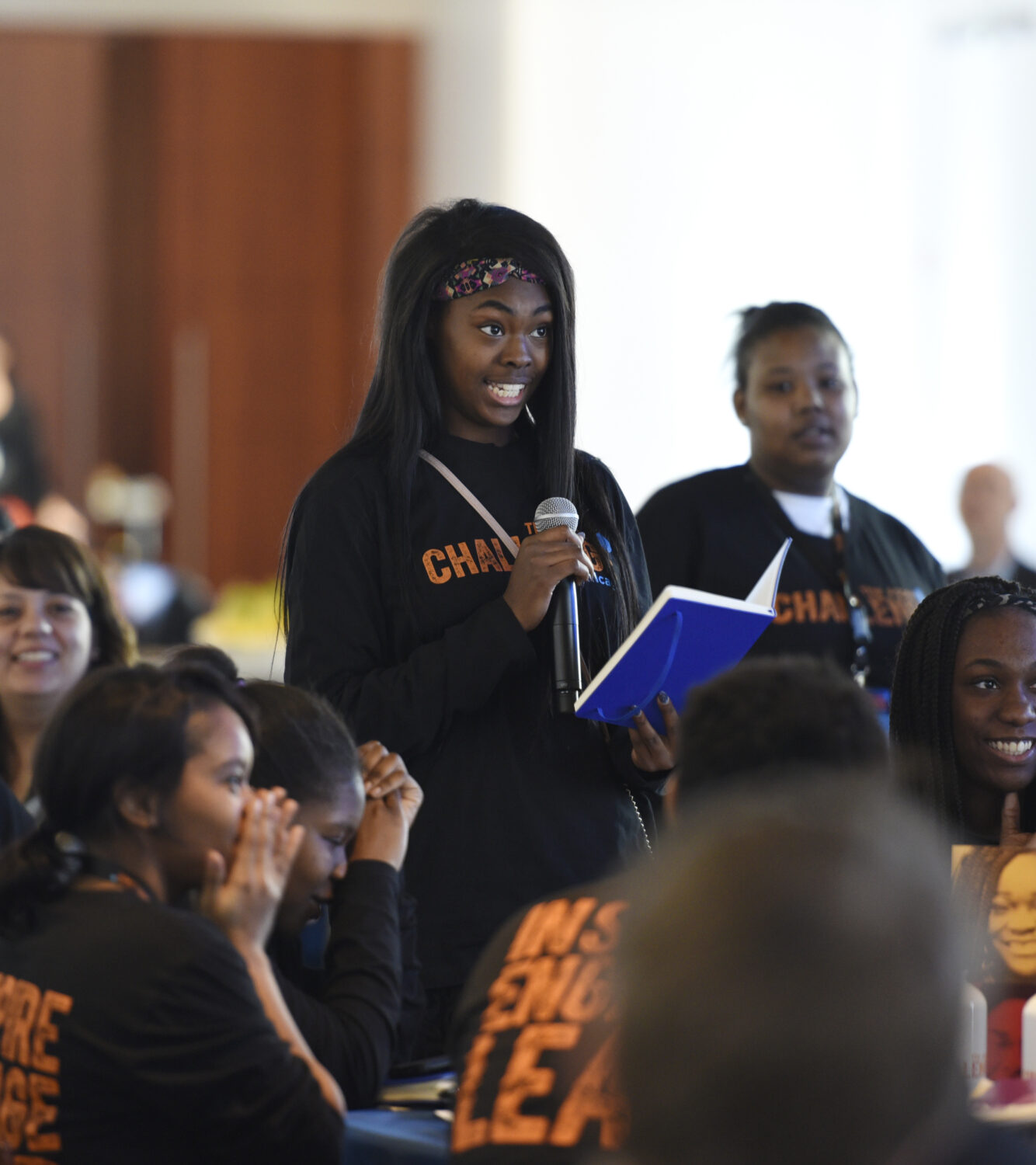 A student, holding a notebook, speaks into a microphone. She is surrounded by a cheering group of young people wearing matching black and orange t-shirts.